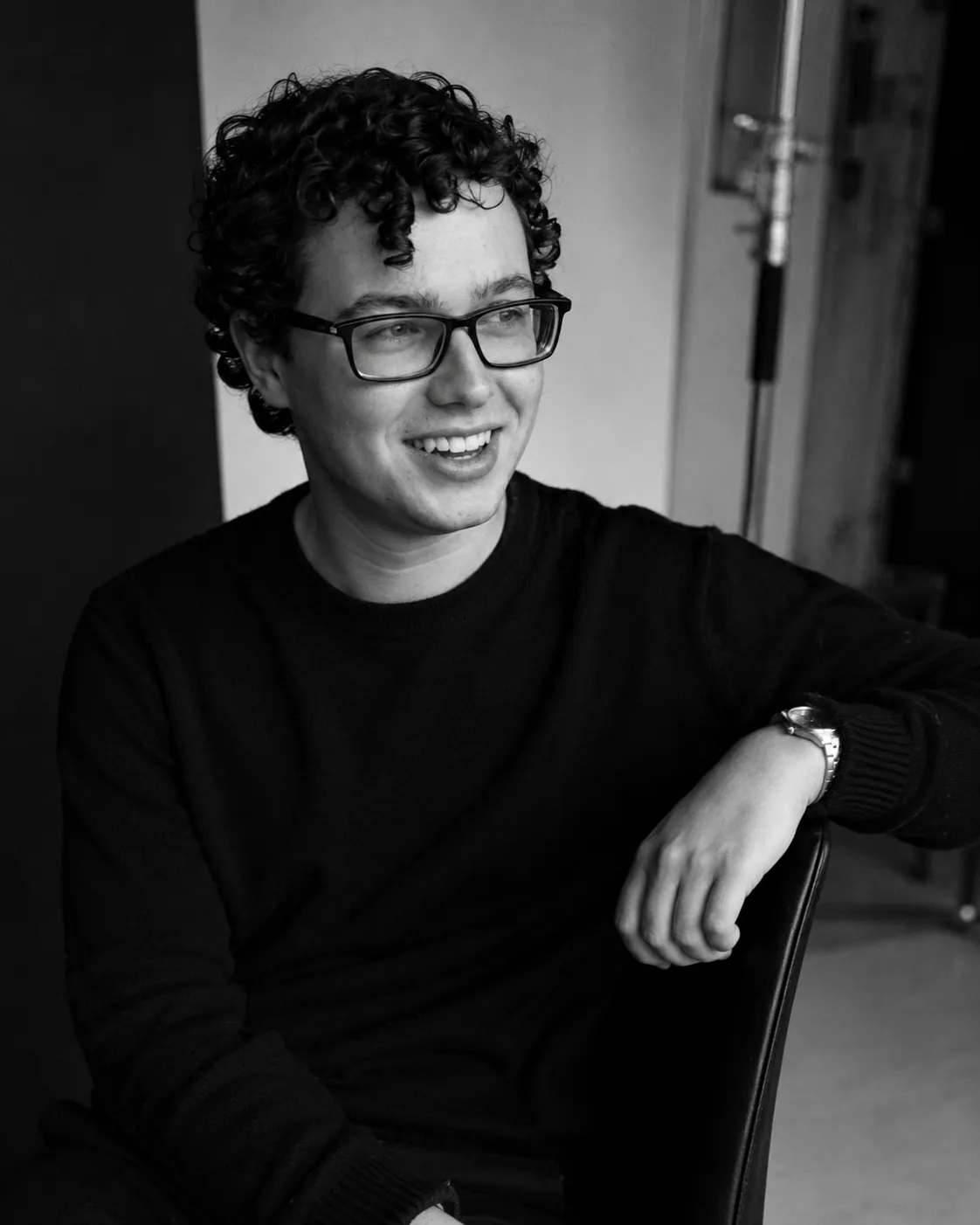 Black-and-white portrait of Matthew Mashford, seated, arm resting on a chair, smiling away from the camera